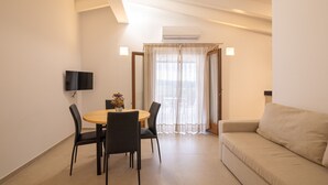 A modern living room with a beige sofa, a round dining table with four chairs, a flat-screen TV mounted on the wall, and a large window with sheer curtains.