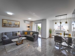 A modern living room with a grey sectional sofa, a coffee table, and a dining area with a glass table and chairs. - Kastel Stari Sunny Downtown Apartment With Terrace (Kastela)