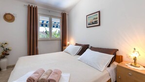 A bedroom with a bed, a nightstand, a window with curtains, a wall-mounted picture, and a towel rack with towels. - Bright and Spacious Apartment With Terrace in Kastel Stafilic (Kastela)