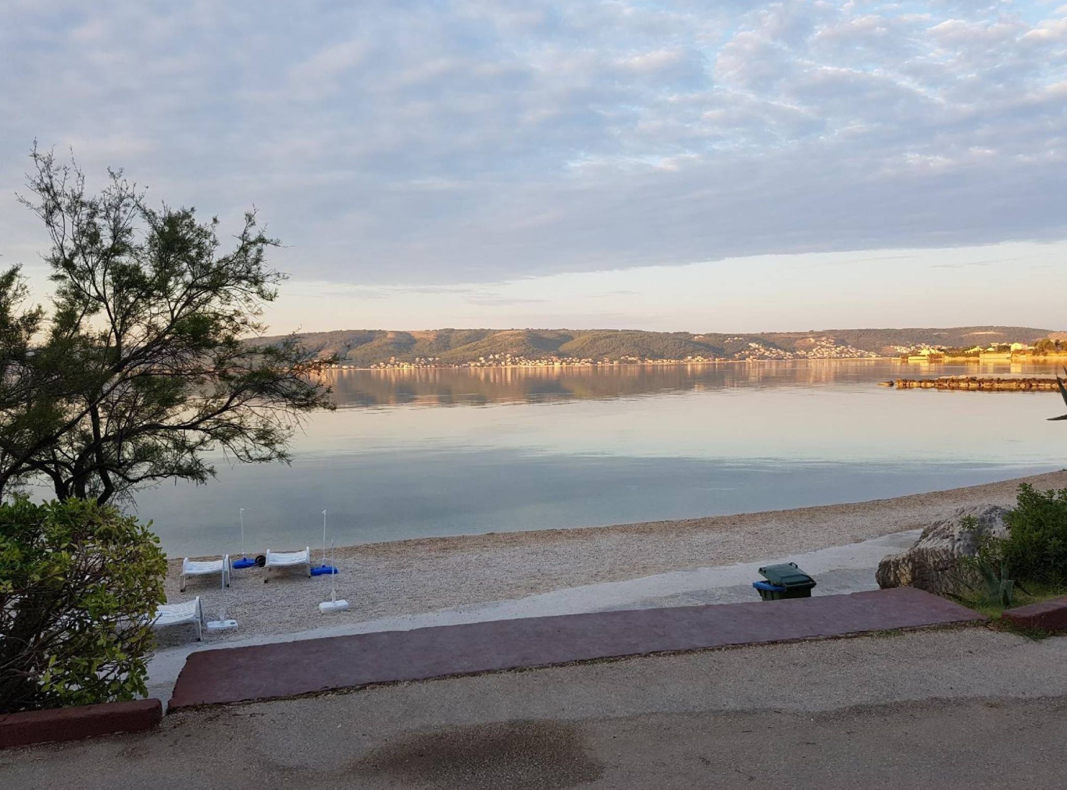 A lakeside view with a pebble beach, a row of white chairs, and a small boat.