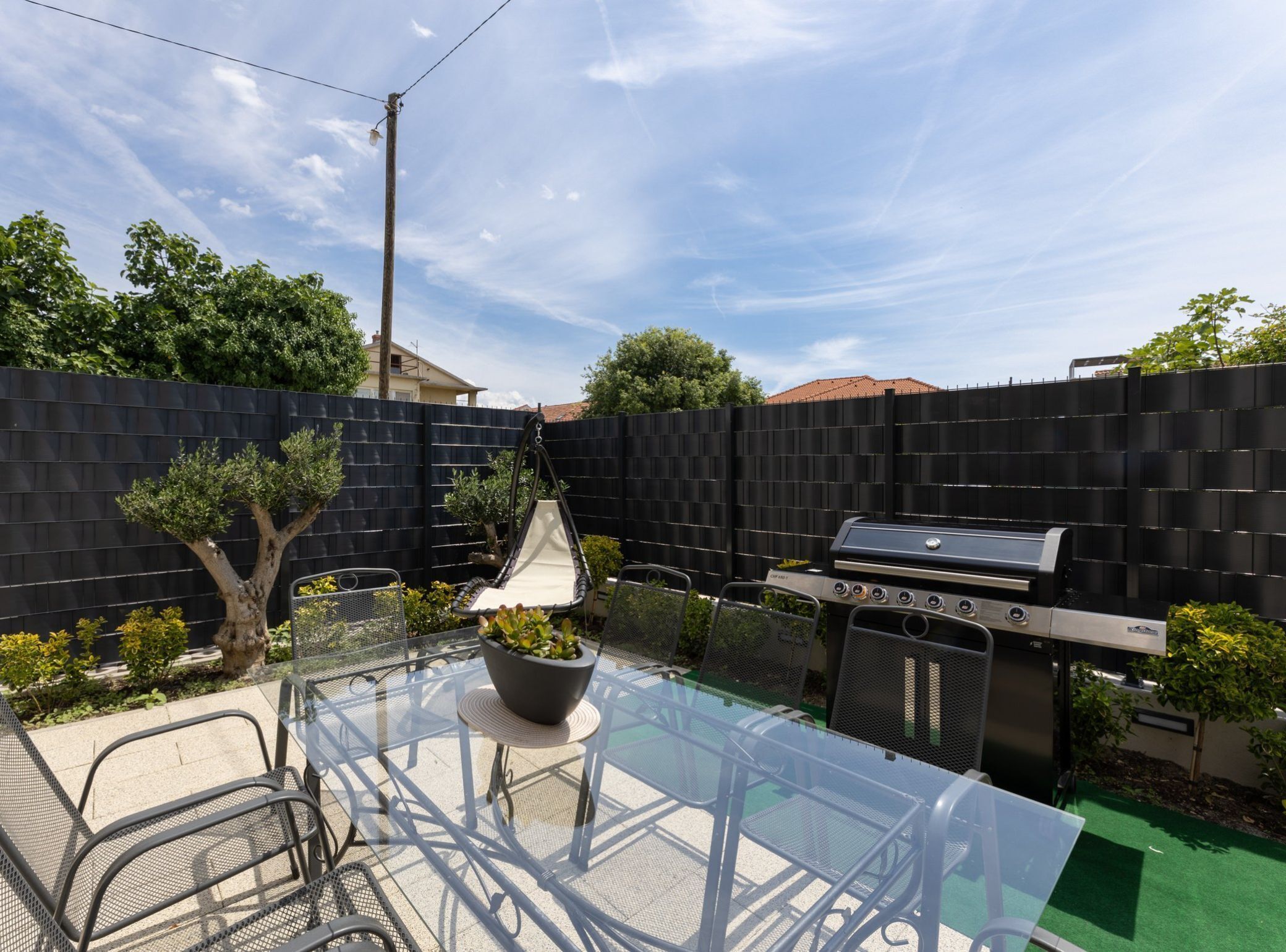 Un giardino attrezzato con un tavolo in vetro, sedie, una poltrona sospesa, un barbecue e un recinto nero.