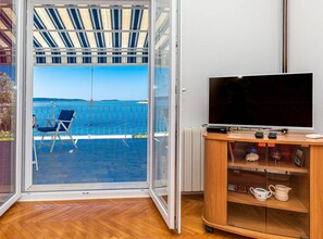 Ein Zimmer mit einer Schiebetür, die auf einen Balkon mit Meerblick führt. Auf dem Balkon befindet sich ein blauer Stuhl, im Zimmer ein Fernseher auf einem Holzboden und eine kleine Ablagefläche mit Dekoration.