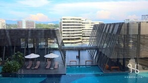 Area piscina sul tetto con sedie a sdraio e ombrelloni, circondata da edifici moderni e un cielo sereno.