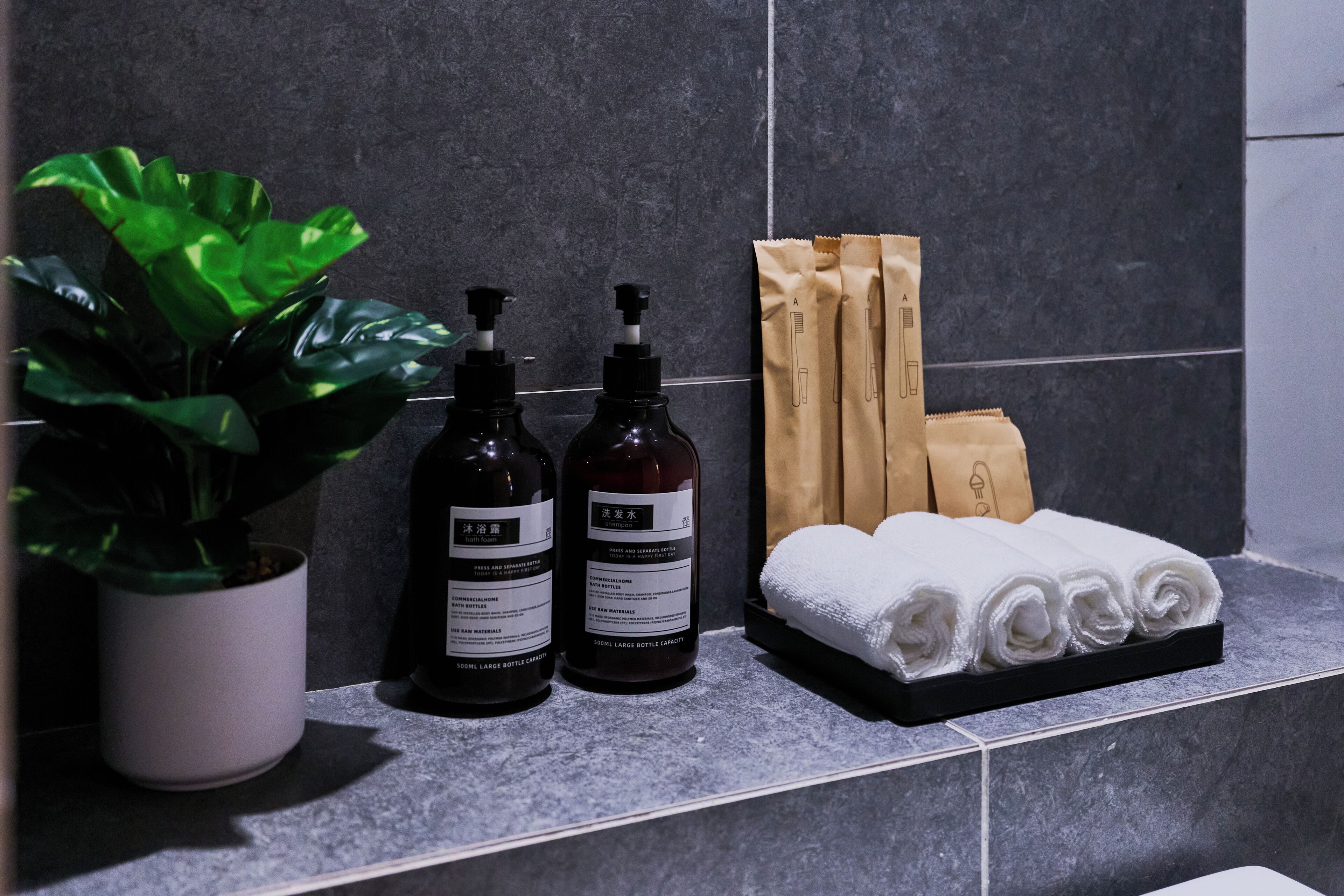 A bathroom counter with a potted plant, two bottles of liquid soap, a towel rack with neatly rolled towels, and a brown paper bag.