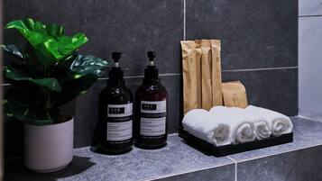 Un meuble-vasque de salle de bain avec une plante en pot, deux bouteilles de savon liquide, un porte-serviettes avec des serviettes soigneusement pliées, et un sac en papier brun.