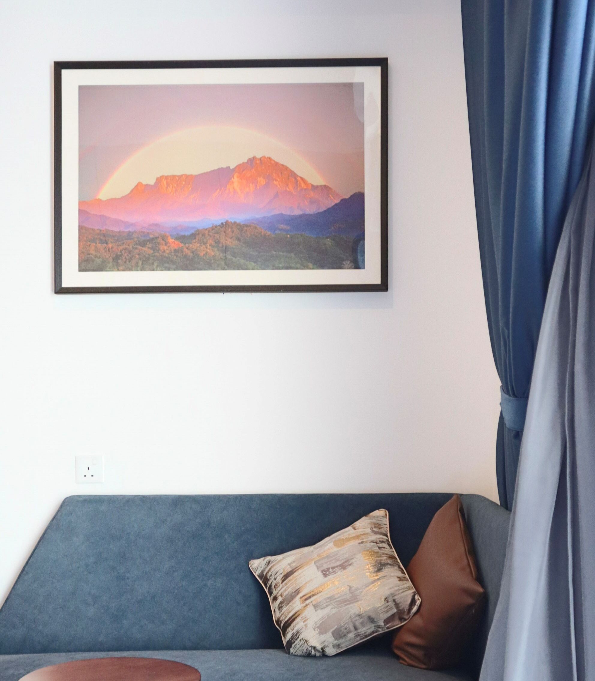 A framed landscape painting of a mountain range with a rainbow on a wall, a blue sofa with a brown pillow, and a blue curtain.