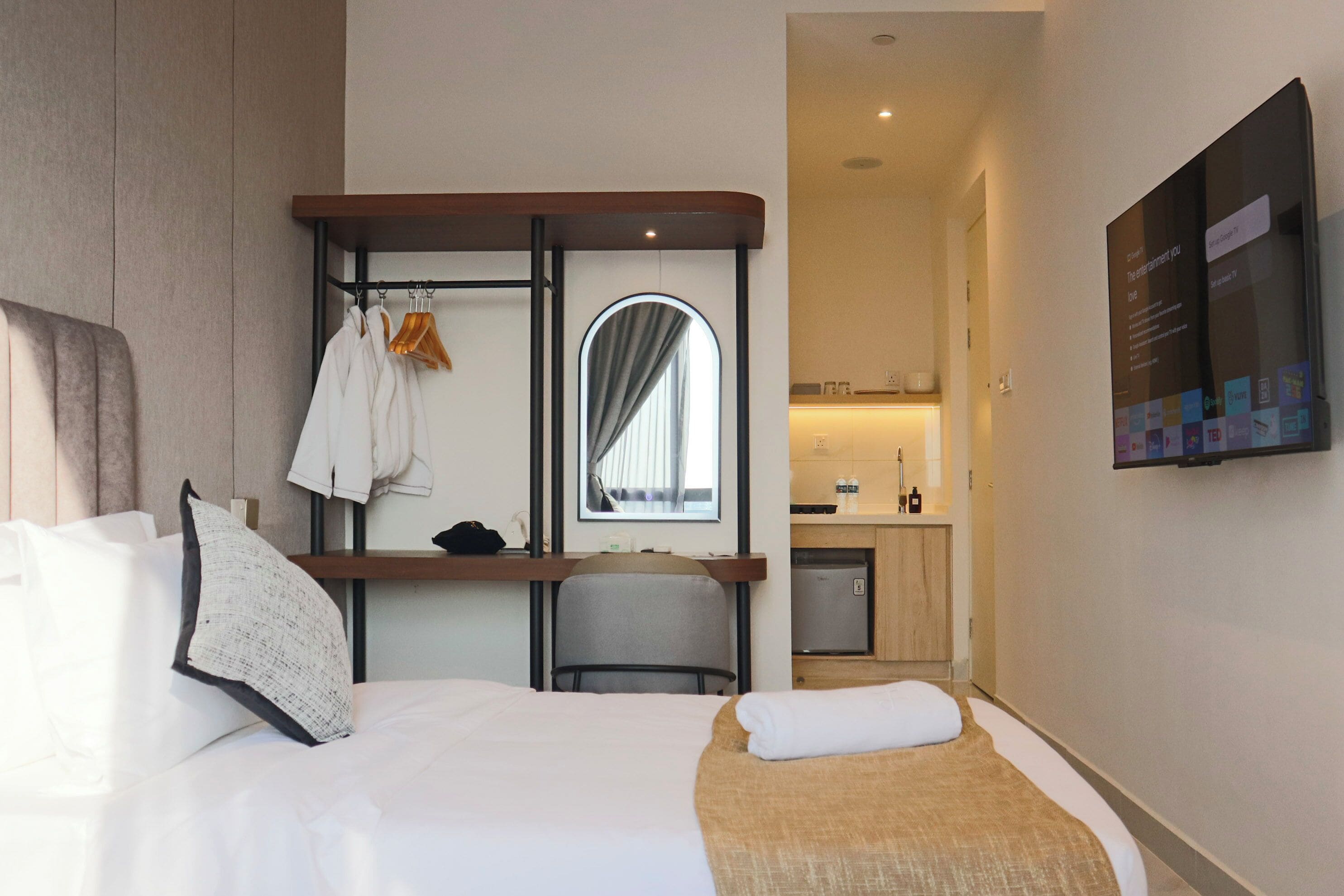 Habitación de hotel con cama, escritorio con silla, espejo, kitchenette pequeña y televisor montado en la pared.