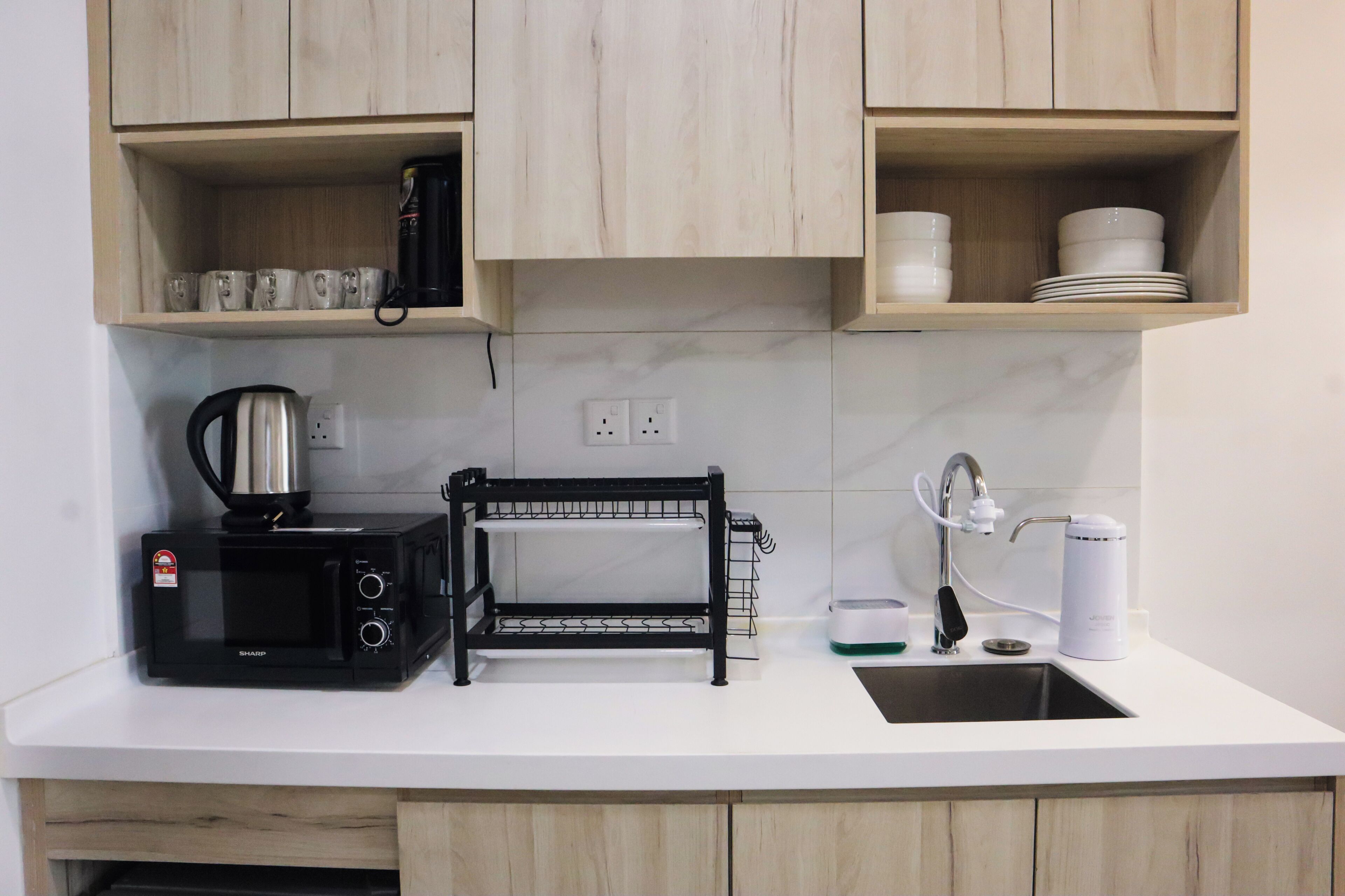 Una cocina con microondas, hervidor de agua y una estantería pequeña. Hay estantes arriba con tazas y platos.
