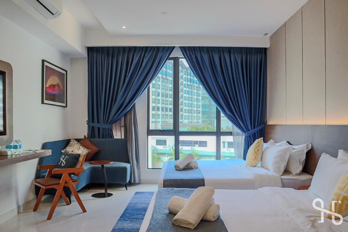 A hotel room with a large bed, a sofa, a desk, and a chair. There is a window with curtains, a painting on the wall, and a view of buildings outside.