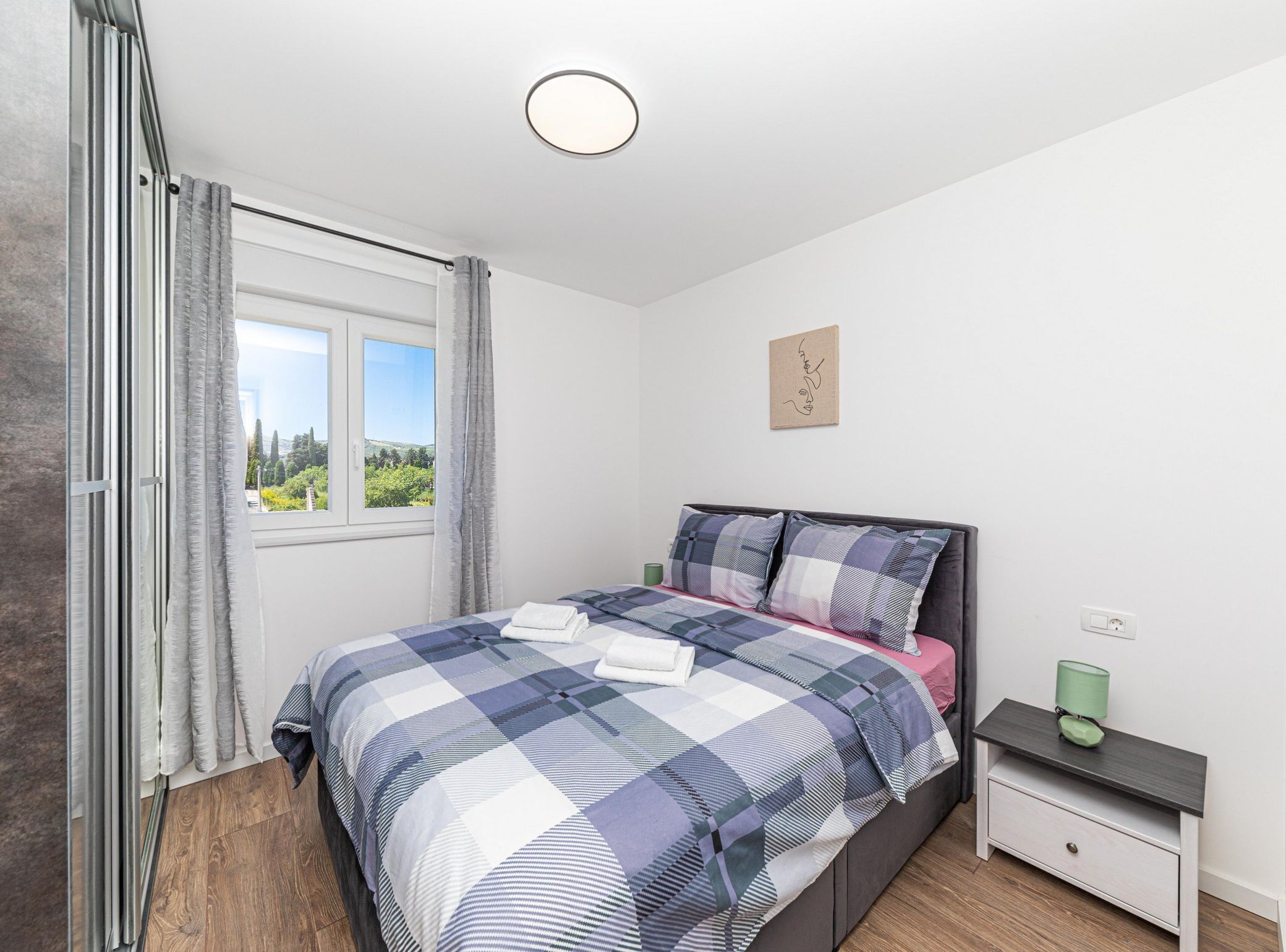Une chambre à coucher avec un lit, une table de chevet, une commode, une fenêtre et une vue sur la verdure à l’extérieur.