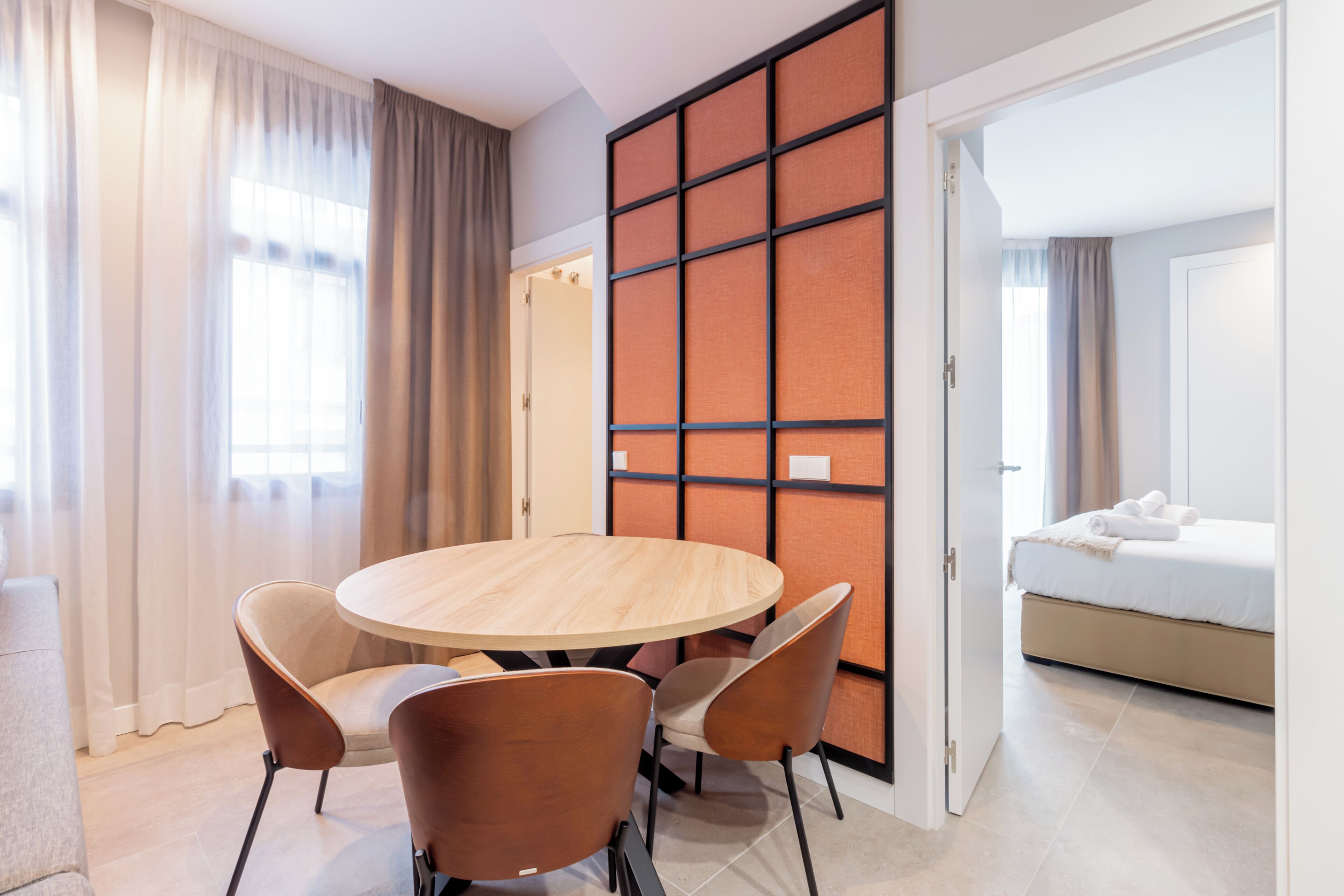 Habitació d'hotel amb una taula rodona de fusta, quatre cadires, un llit amb roba blanca i una porta corredissa amb un disseny de panells.