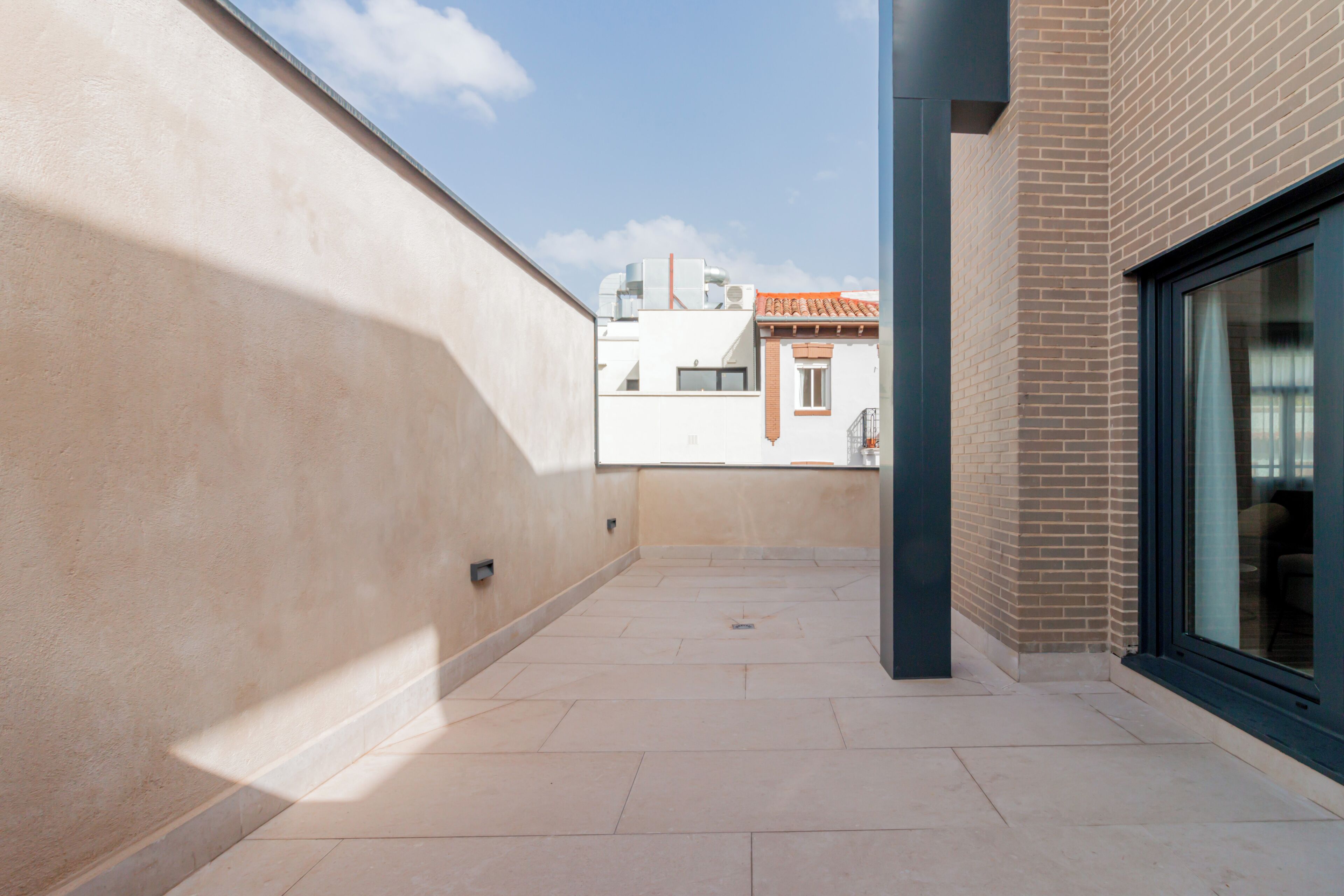 Un estrecho pasillo exterior con suelo de baldosas, pared beige y una puerta de cristal a la derecha.