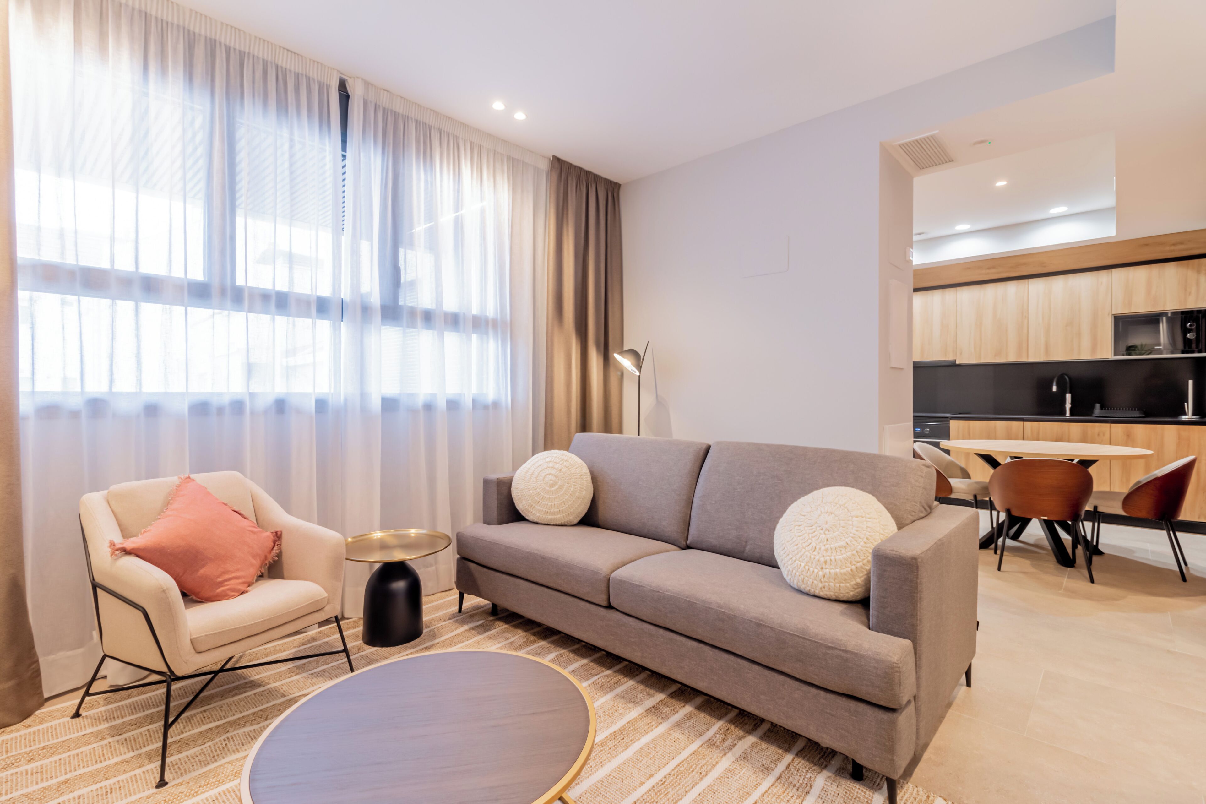 Una sala de estar moderna con un sofá, una mesa de centro redonda y un sillón. Se ve un área de cocina al fondo.