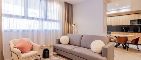 Una sala de estar moderna con un sofá, una mesa de centro redonda y un sillón. Se ve un área de cocina al fondo.