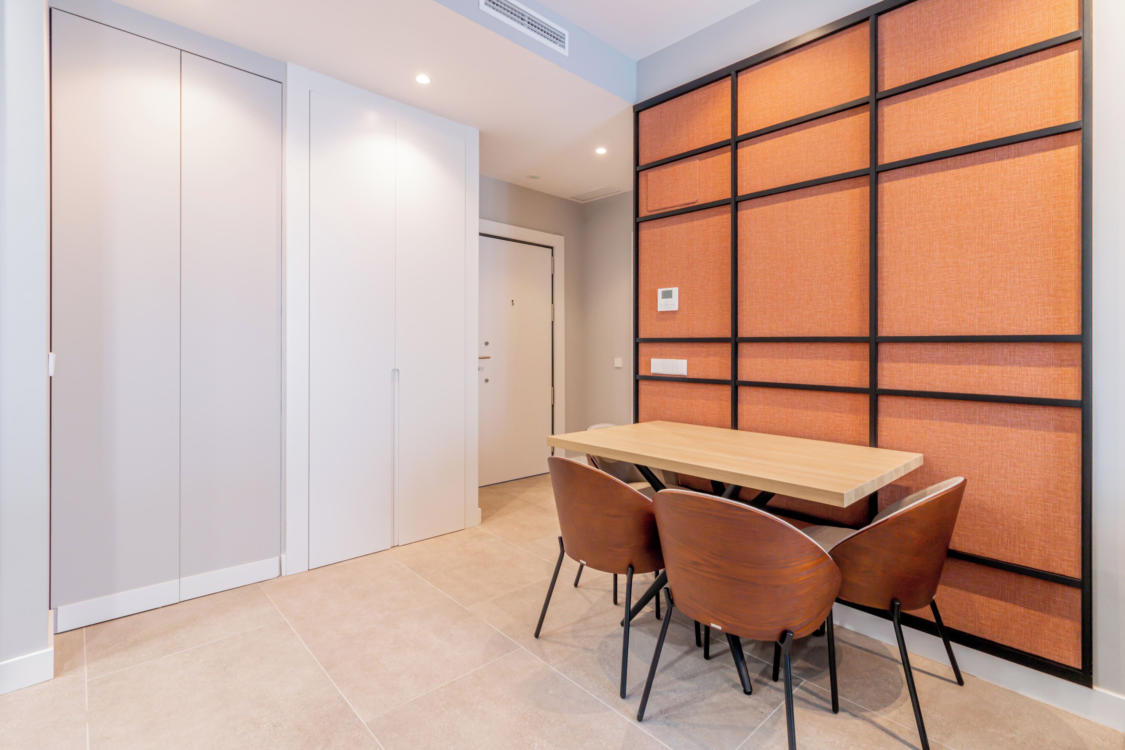 Una habitación moderna con una mesa de madera y cuatro sillas, un armario blanco y una pared divisoria con diseño de cuadrícula.
