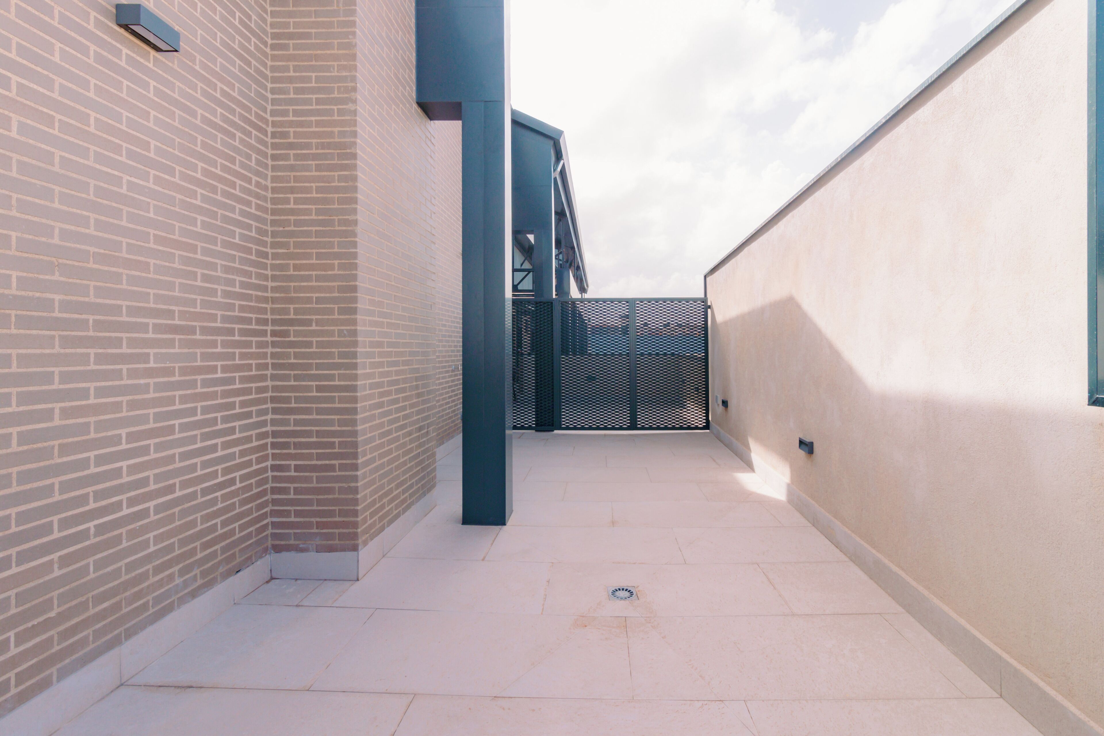 A narrow corridor with a brick wall on one side and a plain wall on the other, leading to a metal gate.