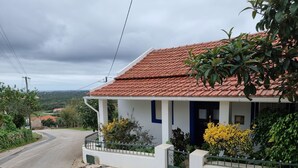 A house with a red-tiled roof, white exterior walls, and a blue door. It has a stone-paved front yard with a metal fence and various plants. - Casa da Ti Alzira - a refuge at the foot of the mountain with a touch of tradition (Leiria)