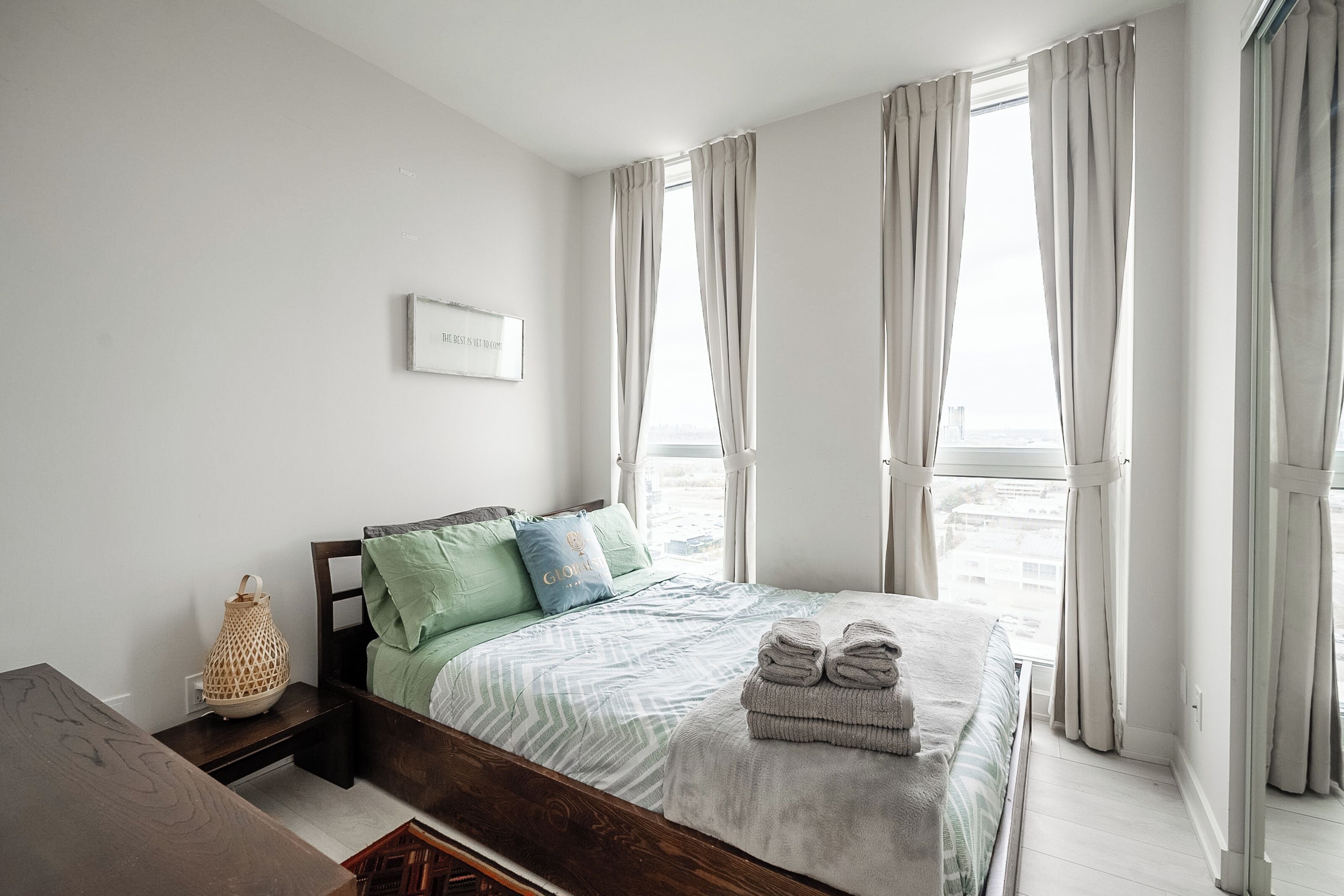 Une chambre à coucher avec un lit, des oreillers, une table de chevet avec une lampe et une fenêtre avec des rideaux.