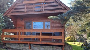Une cabane en bois dotée d’un balcon, entourée d’arbres et d’un mur de pierres.