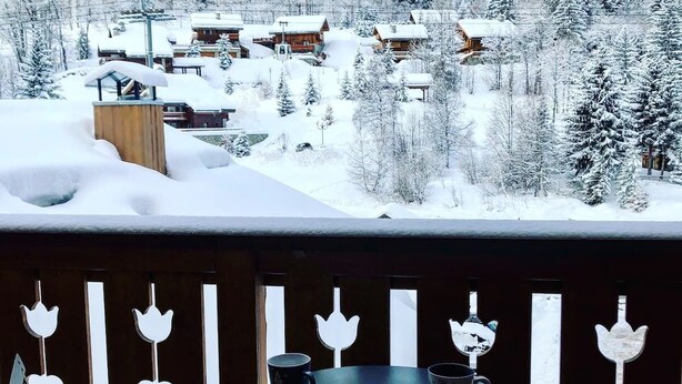 Ein verschneites Dorf mit Holzhäusern und einem Balkon mit dekorativen Metallverzierungen.