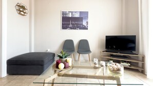 A modern living room with a glass coffee table, two grey chairs, a black sofa, a flat-screen TV, and a cityscape picture on the wall.