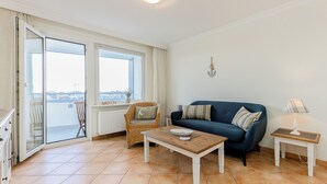 A living room with a blue sofa, a wooden coffee table, a wicker chair, and a view through a glass door. - WindSpiel - House Ankerlicht - Westerland (Westerland)