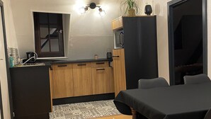 Een knusse keuken met houten kastjes, een zwarte koelkast en een magnetron. Er is een eettafel met stoelen, een klein vloerkleed en een plant in pot.