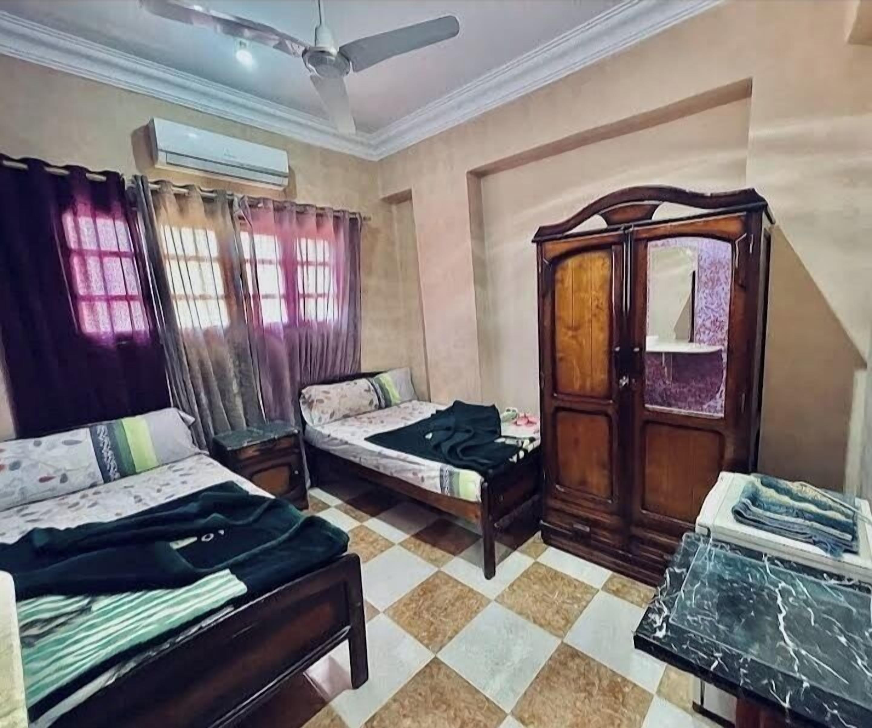 Quarto com duas camas de solteiro, guarda-roupa de madeira, ventoinha no teto e piso ladrilhado.