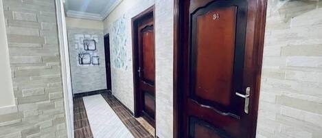 Un pasillo con dos puertas de madera, cada una con un número. Las paredes tienen un diseño que imita la piedra, y el suelo está cubierto de baldosas.