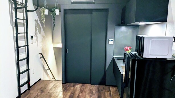 A compact room with a dark door, a small kitchenette with a microwave, a black refrigerator, and a wooden floor.