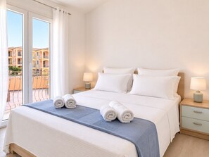 Ein Hotelzimmer mit einem großen Bett, zwei Nachttischen mit Lampen, einem Balkon mit Blick auf Gebäude und zwei zusammengerollte Handtücher auf dem Bett.