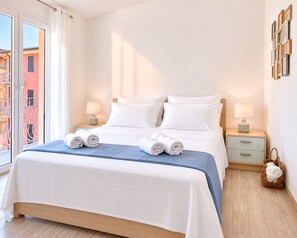 A hotel room with a bed, two bedside tables, a window with curtains, and a view of buildings outside. - Chris House Key to Villas (Sardegna)