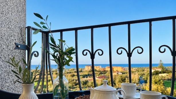 Un balcone con vista sul mare, un tavolo con teiera, tazze, una ciotola di frutta secca e un mescolino per il miele.