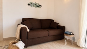 Un divano marrone con una coperta bianca, un cesto intrecciato con un asciugamano bianco e un tavolino con un libro e un oggetto decorativo.