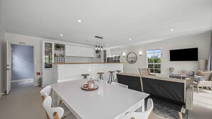 Uma cozinha moderna com armários brancos, uma sala de jantar com mesa e cadeiras brancas, e uma sala de estar com sofá e televisão.