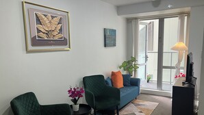 Un salon avec un canapé bleu, des fauteuils verts, une petite table sur laquelle est posée une plante, une œuvre d’art encadrée et un téléviseur posé sur un meuble.