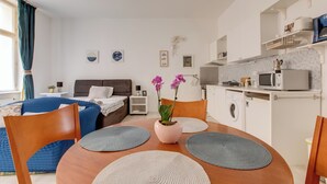 A modern hotel room with a wooden dining table, two chairs, a potted orchid, and a kitchen area with a microwave and washing machine.