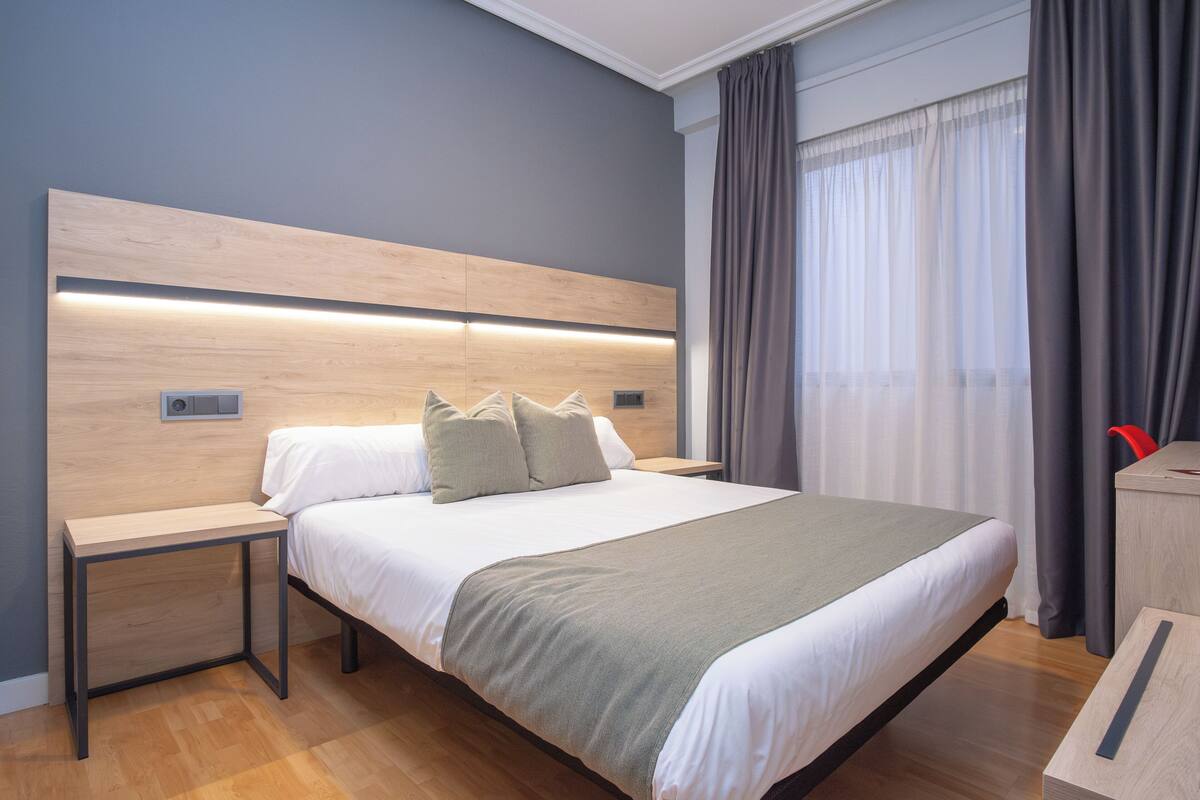 A modern hotel room with a wooden headboard, a large bed with a green throw, a bedside table, and a nightstand.