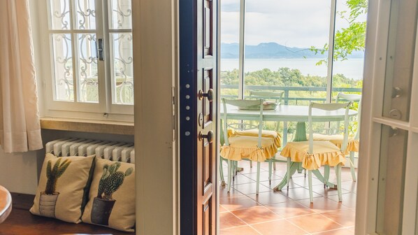 An open door to a balcony with a table and chairs, a view of a body of water, and a cactus plant on a bench. - Villa Castlegrace Lake Garda - Italian Homing (Padenghe sul Garda)