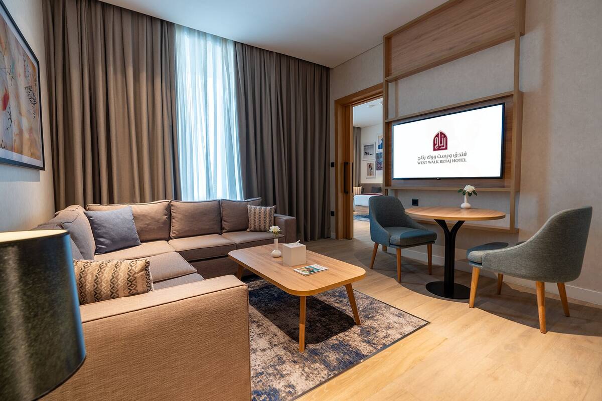 A modern hotel room with a sofa, a coffee table, and two chairs. There is a large window with curtains, a flat-screen TV mounted on the wall, and a framed picture on the wall.