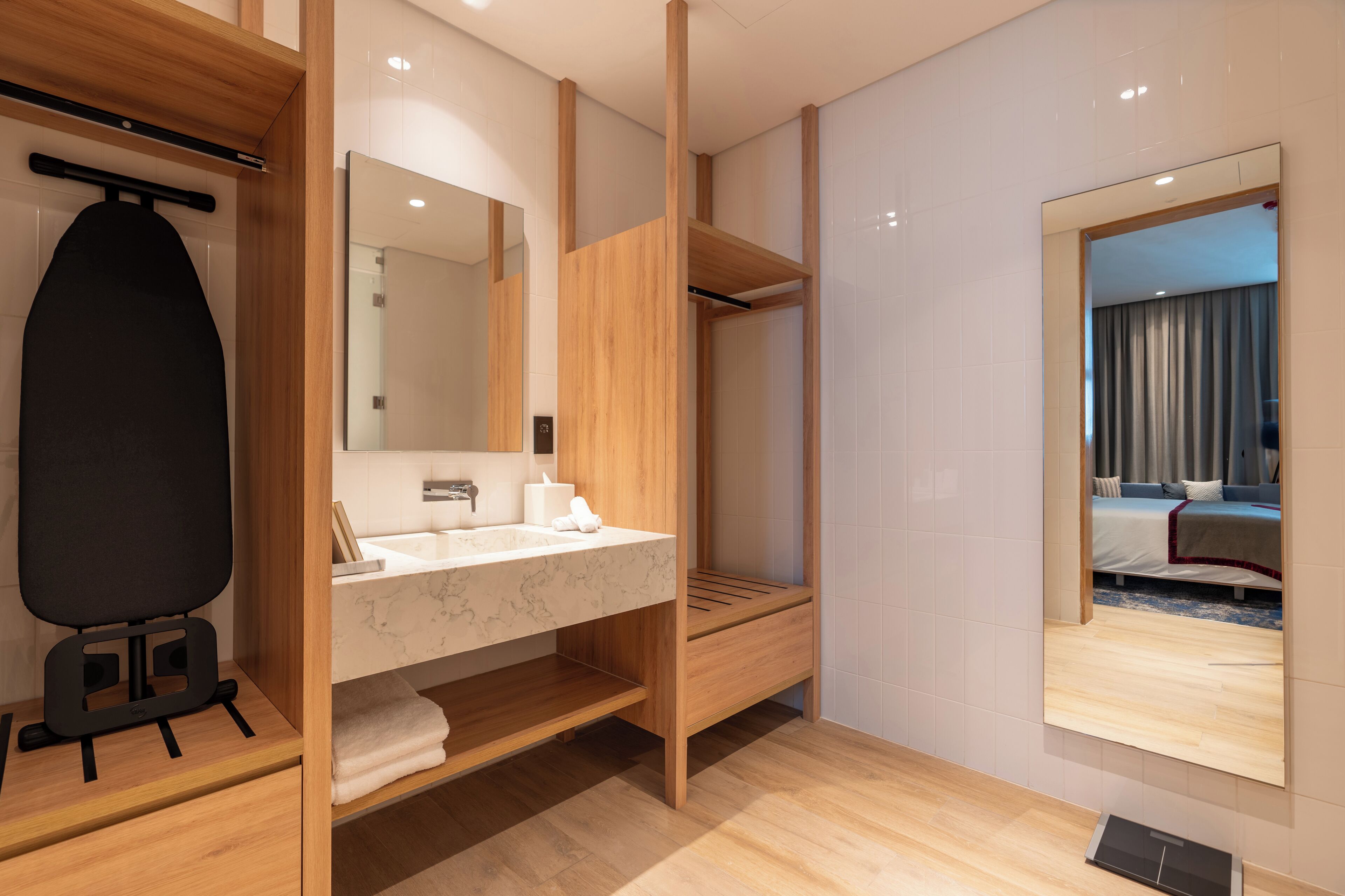 Een moderne badkamer met een houten wastafelmeubel, een grote spiegel en uitzicht op een slaapkamer.