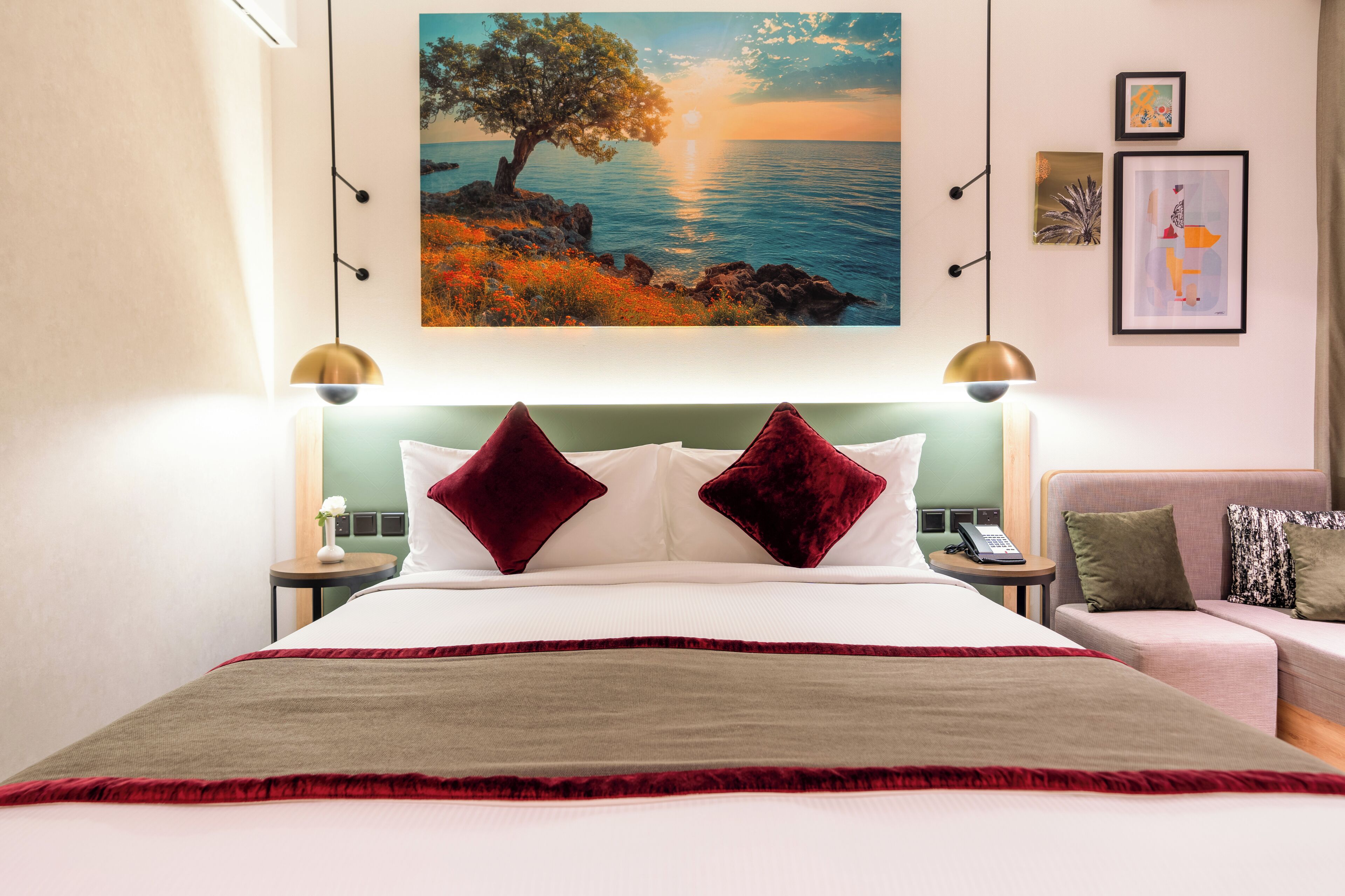 A hotel room with a large bed, two red pillows, a painting of a tree by the sea, and a small table with a phone.