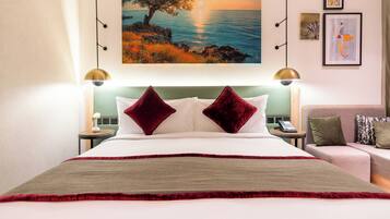 A hotel room with a large bed, two red pillows, a painting of a tree by the sea, and a small table with a phone.
