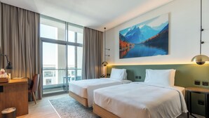 Ein Hotelzimmer mit zwei Betten, einem Schreibtisch, einem Stuhl, einem kleinen Tisch und einem großen Fenster mit Vorhängen.