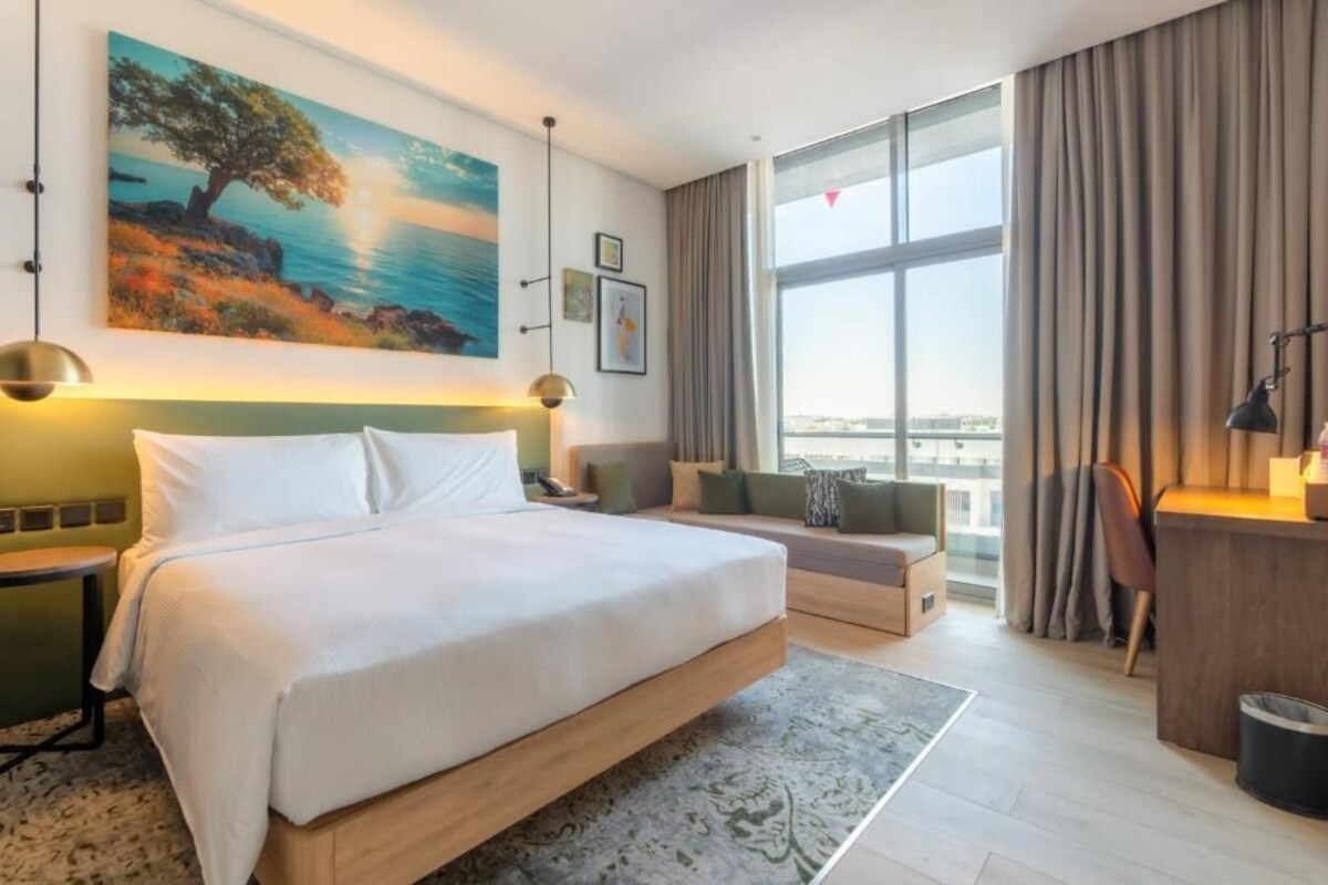 Ett hotellrum med en stor säng, ett skrivbord, en stol, utsikt över staden och en tavla på väggen.