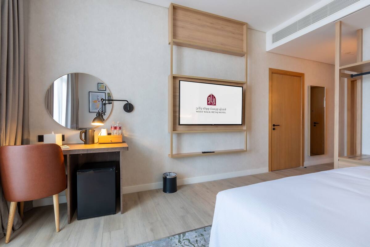 A hotel room with a bed, a desk with a computer, a chair, a mirror, a TV, and a door.