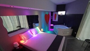 A modern hotel room with a large bed, a sitting area with a chair, a small table, a wall-mounted TV, and a bathtub labeled 'LIVING B&B'. - Living (Casoria)