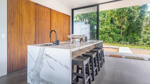 Cocina moderna con una isla de mármol, panel de pared de madera y un amplio ventanal corredero que da a un jardín.
