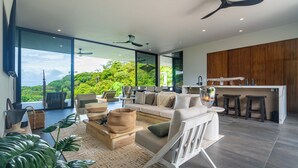 Una sala de estar moderna con un amplio ventanal, un ventilador de techo de madera y una zona de cocina con taburetes.