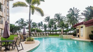 Ein Schwimmbecken mit Liegestühlen und Sonnenschirmen, umgeben von Palmen und einem Gebäude mit Balkonen.